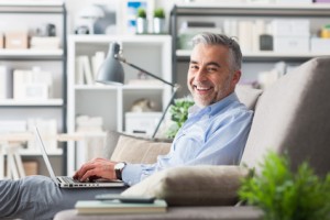 Cheerful confident businessman at home sitting on the couch and working with a laptop, he is smiling at camera