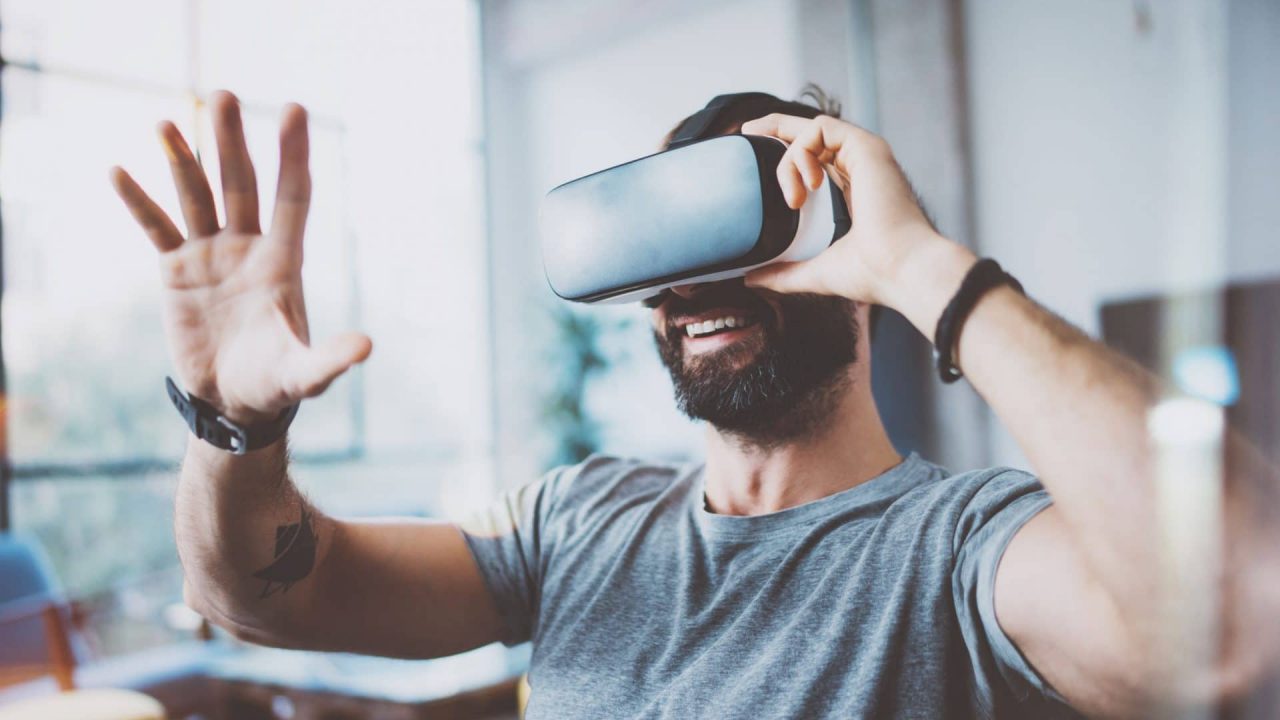 67840133 - young bearded man wearing virtual reality glasses in modern interior design coworking studio. smartphone using with vr goggles headset. horizontal,flares effect, blurred background